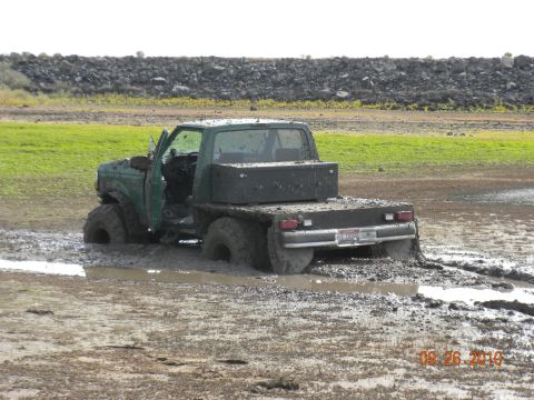 1989 Ford Bronco - This green flatbed truck is deeply stuck in mud, with only its rear visible.