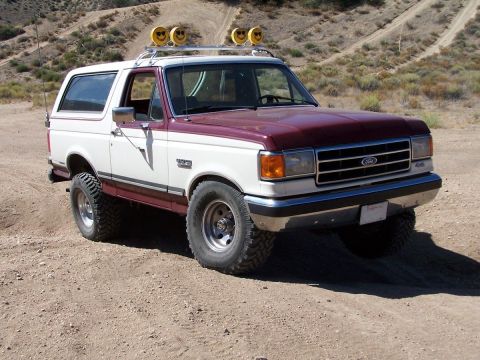 1990 Ford Bronco - A two-tone 1990 Ford Bronco with a roof rack and off-road tires is parked on a dusty, unpaved road in a dry, hilly area.