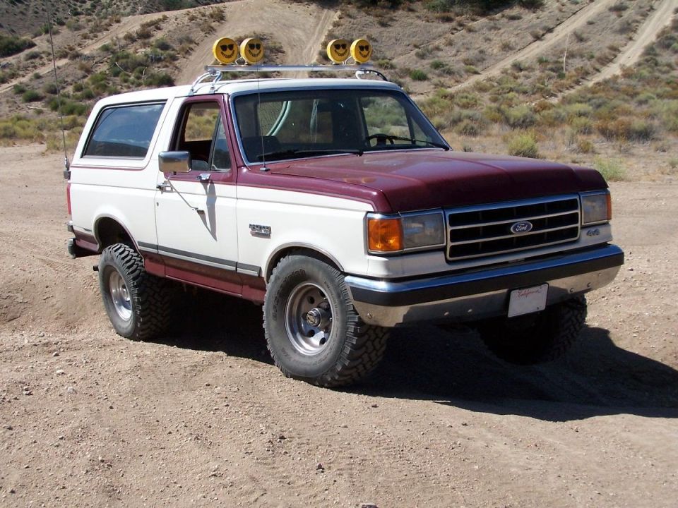 White and maroon 1990 Ford Bronco with roof rack and off-road tires parked on a dirt road