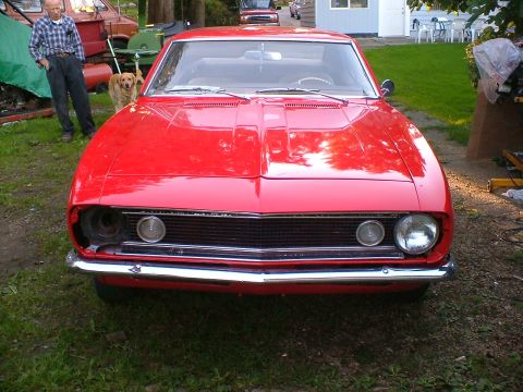 1989 Ford Bronco - A red 1969 Chevrolet Camaro is parked outdoors with a man and a dog nearby.