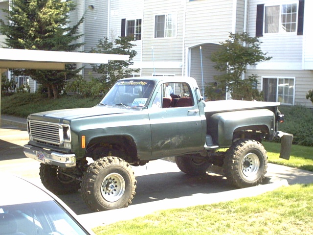 Lifted dark green stepside pickup with white top and big mud tires, 3/4 front view