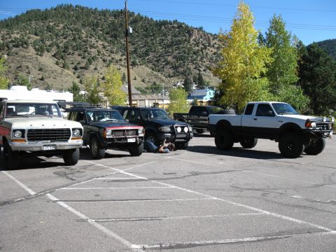 1978 Ford Bronco - Several modified trucks and SUVs are parked in a lot with a mountainous backdrop.