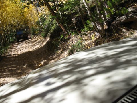 1978 Ford Bronco - A dark SUV navigates a winding dirt road surrounded by trees with yellowing leaves.