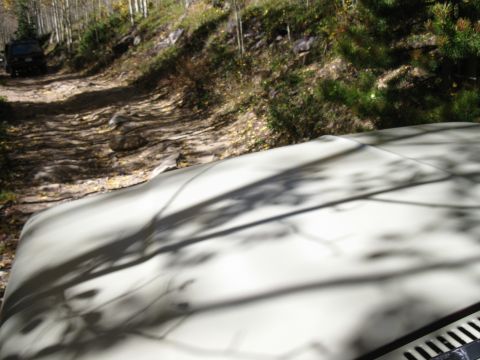 1978 Ford Bronco - A white vehicle's hood is in the foreground, with a dark SUV driving on a dirt road ahead.
