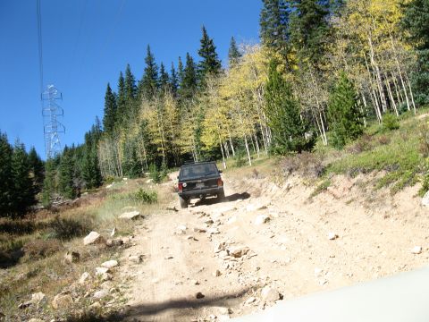 1978 Ford Bronco - A black SUV navigates a rough dirt road through a wooded mountainside under a clear blue sky.