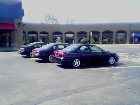 1999 Ford Taurus - Three dark sedans are parked in a lot in front of a strip mall.