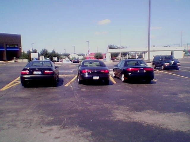 Four vehicles parked in a lot: three dark sedans and a dark SUV. The sedans are in the foreground, viewed from the rear