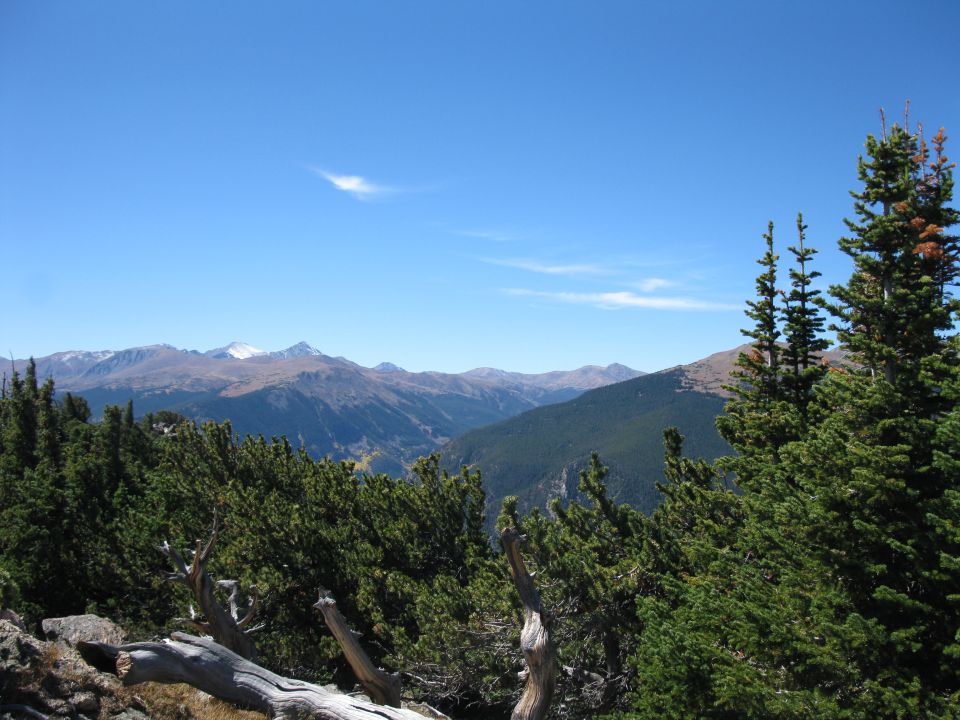 Mountain valley with snow-capped peaks and pine trees in the foreground. Watermark reads SUPERMOTORS.NET