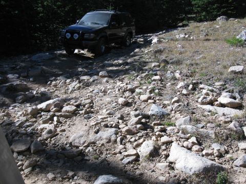 1978 Ford Bronco - SUV driving on a rocky mountain road with trees in the background.