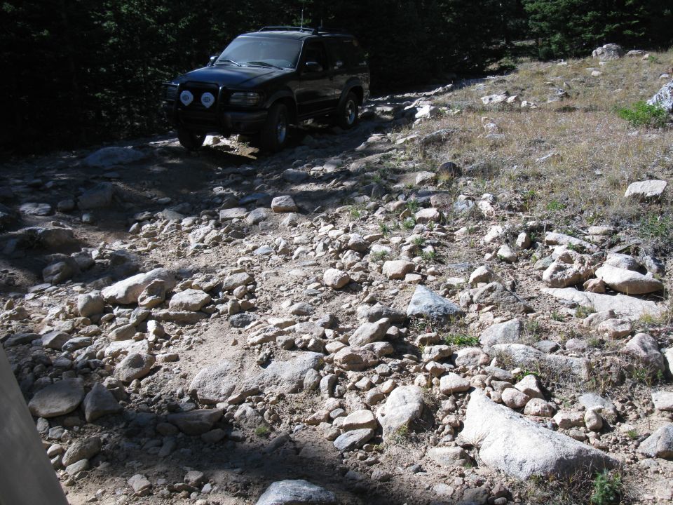 Black SUV navigates a rough, rocky dirt road with trees in the background