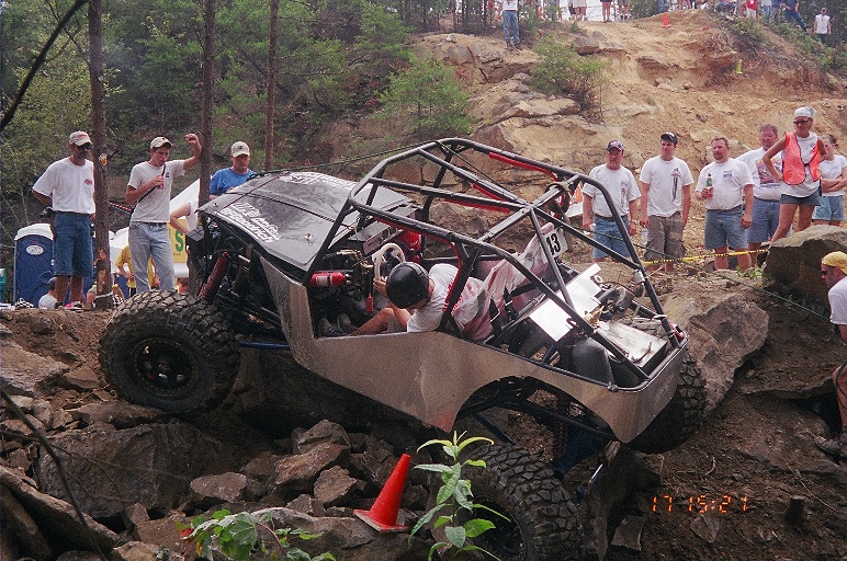 Number 13 rock buggy tipped on rocks with driver leaning out, spectators in shirts