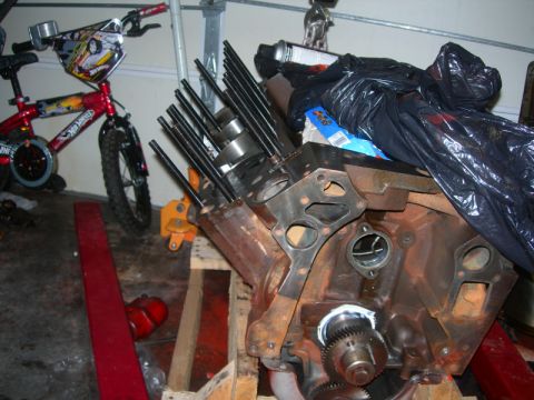 1984 Ford Bronco - Engine block with pushrods and gasket, resting on wooden supports, with a bicycle in the background.