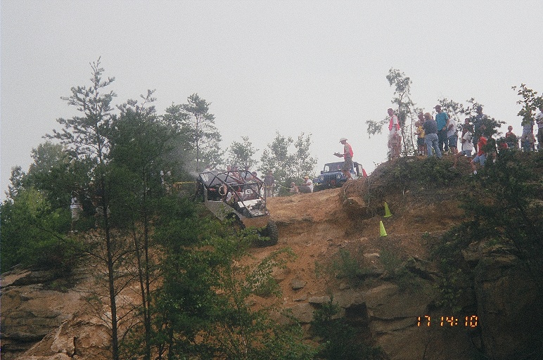 Distant rock buggy cresting a hazy rocky hilltop ridge lined with spectators