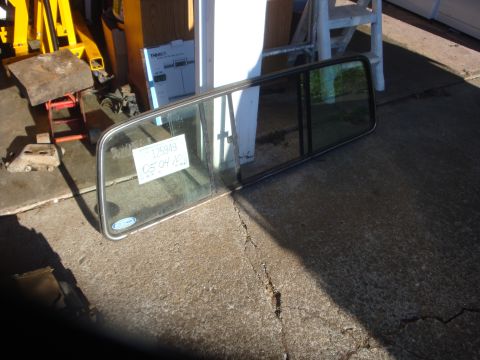 1989 Ford Bronco - A three-panel sliding rear cab window assembly in its metal frame with a blue Ford oval decal, laid flat on a concrete garage floor. A hand…
