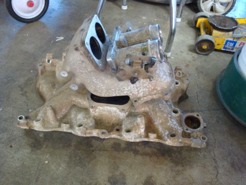 1989 Ford Bronco - An engine intake manifold, likely for a V8, is shown with dual carburetors and throttle linkage, resting on a concrete floor.