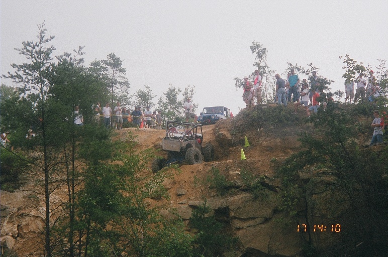Tube-frame rock buggy climbing rocky ledge with crowd of spectators on hilltop above