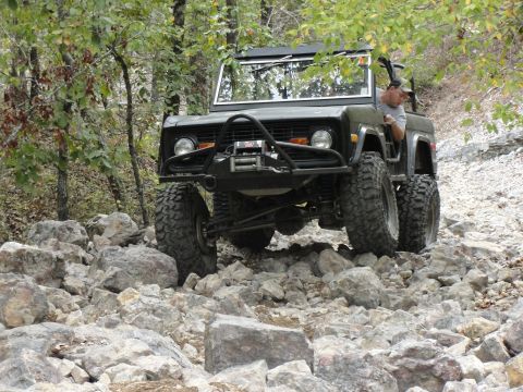 1967 Ford Mustang - A man drives a lifted Ford Bronco over a challenging, rocky terrain surrounded by trees.