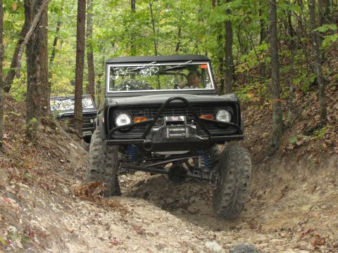 1967 Ford Mustang - Two Ford Broncos navigate a wooded off-road trail with steep inclines and loose dirt.
