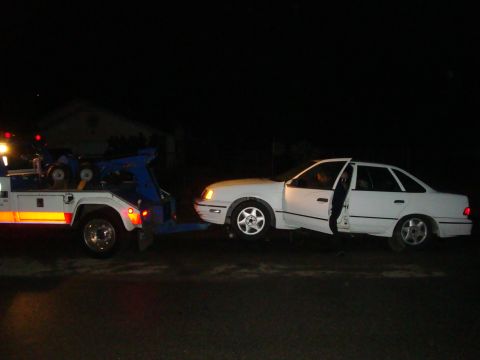 1990 Ford Taurus - A white 1990 Ford Taurus is being towed by a tow truck at night with the driver's door open.