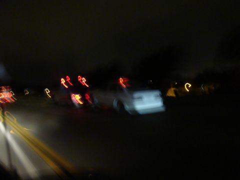 1990 Ford Taurus - Motion blur captures a white sedan on a dark road at night, with streaks of red taillights from other cars visible.