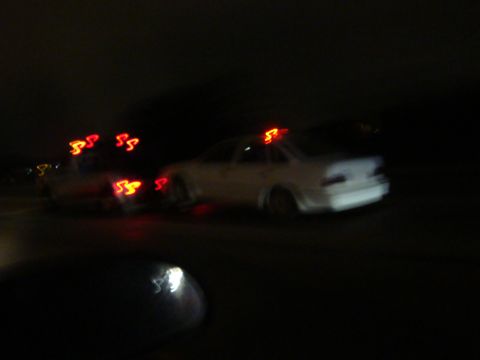 1990 Ford Taurus - The photo shows two cars on the road at night, with the rear car being white and illuminated by its red roof lights.