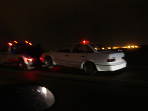1990 Ford Taurus - A white sedan with flashing red lights on its roof is being towed by a truck on a dark road.