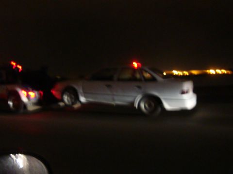 1990 Ford Taurus - A white 1990 Ford Taurus police car with its emergency lights activated is seen from the side at night.