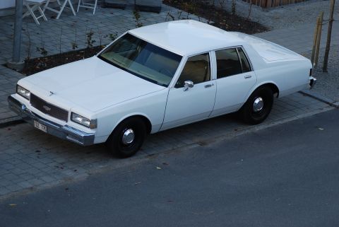 1998 Ford Crown Victoria - This white sedan, likely from the 1980s, is parked on a paved area next to a street. The license plate is visible.