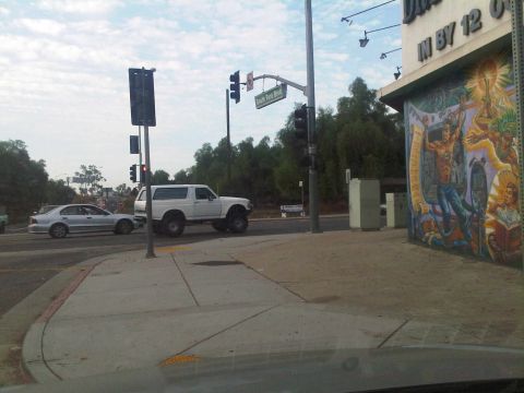 1986 Ford Bronco - A white SUV with lifted suspension and large tires is seen at a street intersection next to a silver sedan.