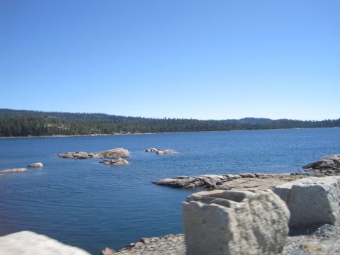 1989 Ford Bronco - A wide shot of a tranquil lake dotted with rocks, bordered by a dense forest under a bright blue sky.