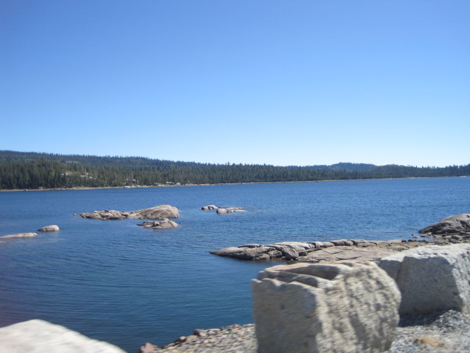Lake with scattered rocks and a distant tree line under a clear blue sky