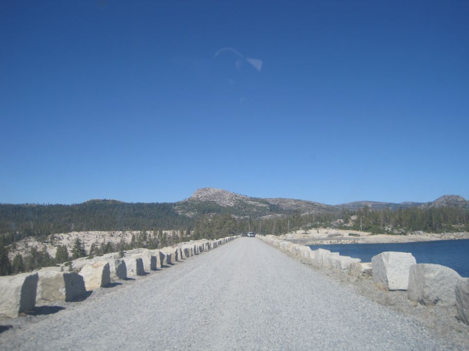 Gravel road leads through a mountain pass next to a blue lake under a clear sky