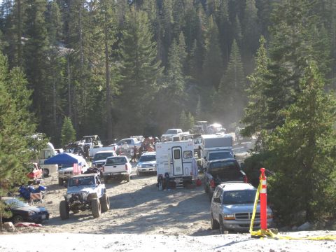 1989 Ford Bronco - A gathering of vehicles, including trucks, SUVs, and campers, are parked on a dirt road surrounded by trees.