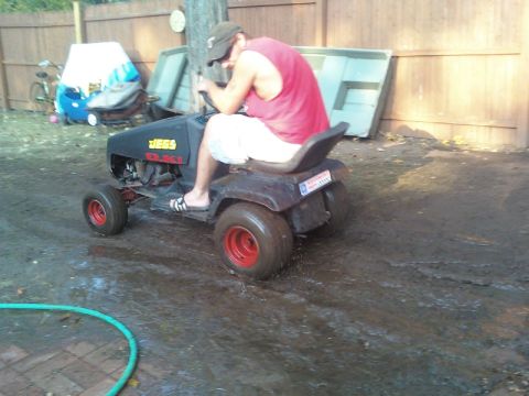 1989 Ford Bronco - A person sits on a modified lawn mower with red wheels and a JEGS logo, parked on a muddy surface.