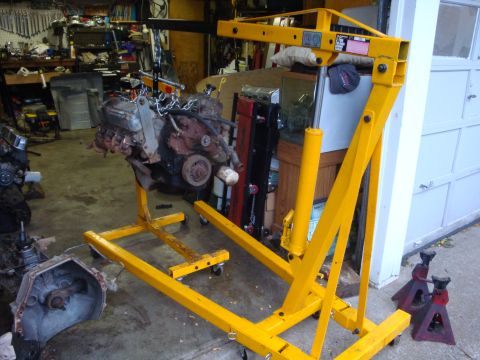 1989 Ford Bronco - An engine is suspended by a yellow engine hoist in a cluttered garage environment.