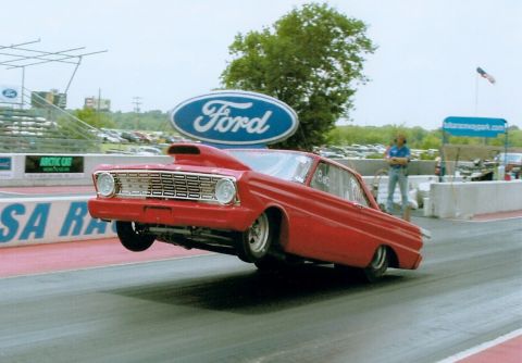 1963 Ford Fairlane - A red drag car lifts its front wheels off the ground during a race at a track with a large Ford sign.