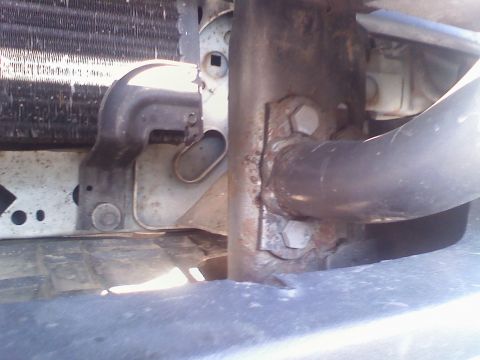1996 Ford Bronco - Close-up under the open hood at the front of a truck, showing the radiator core support and a surface-rusted frame horn with two large hex-…