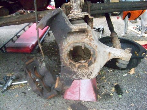 1989 Ford Bronco - Close-up of a rusty front axle spindle and brake assembly, supported by a red jack stand, with tools scattered around.