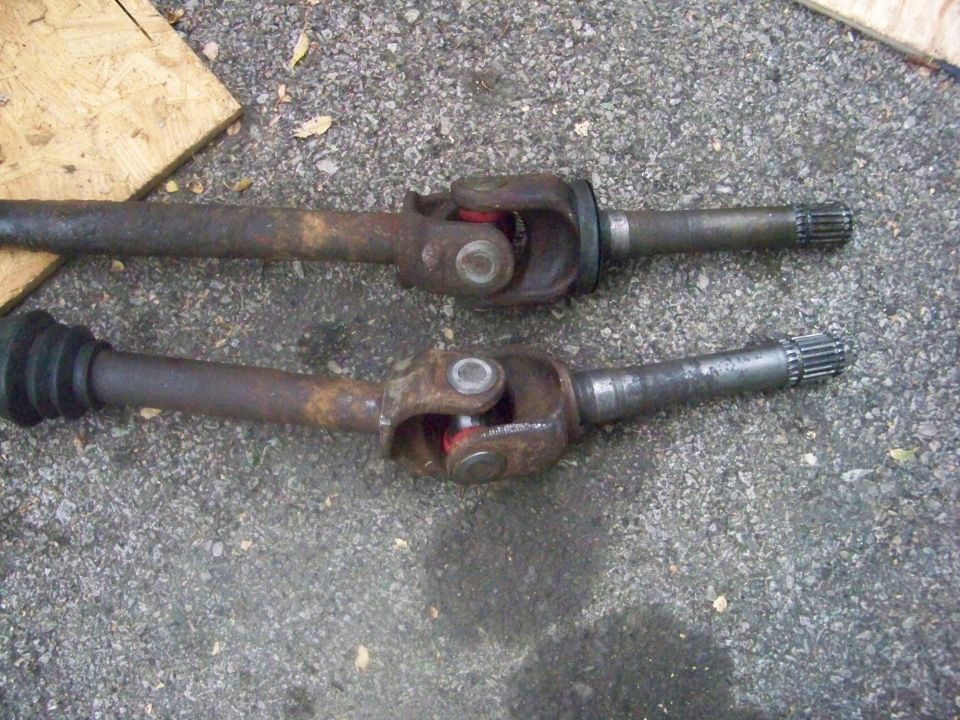 Two rusty driveshafts with U-joints and splined ends, lying on gravel