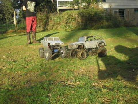 1994 Ford Escort - Two remote-controlled monster trucks are parked on a grassy lawn next to a person.
