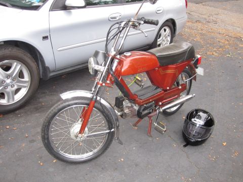 1981 Garelli Rally Sport - A red moped is parked next to a silver car, with a black helmet resting on the asphalt.