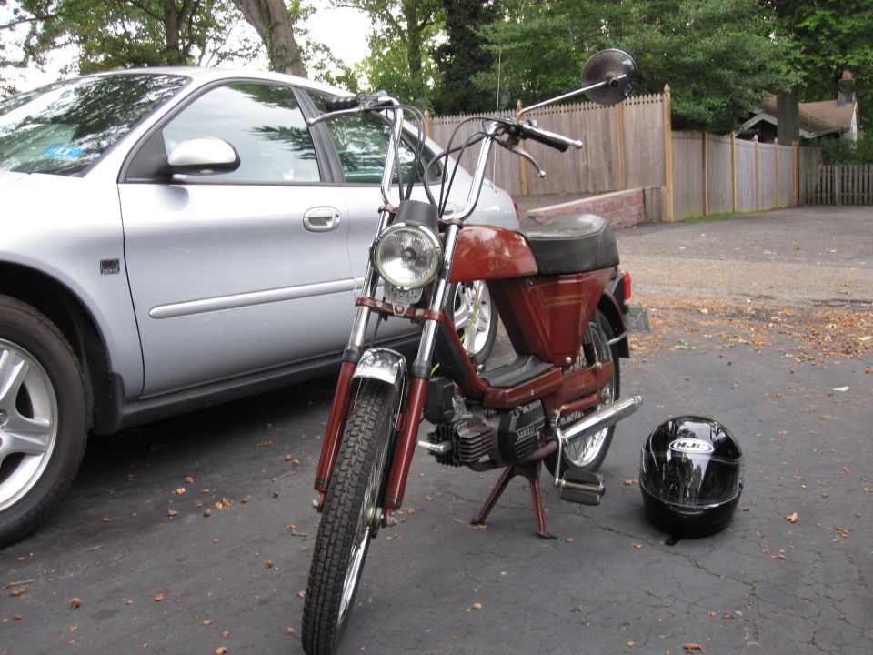Red moped parked next to a silver sedan, with a black helmet on the ground