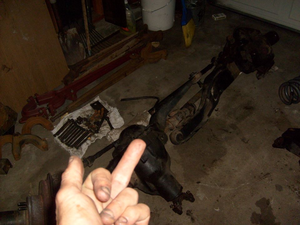 Vehicle axle and suspension parts laid out on a garage floor. A hand is in the foreground