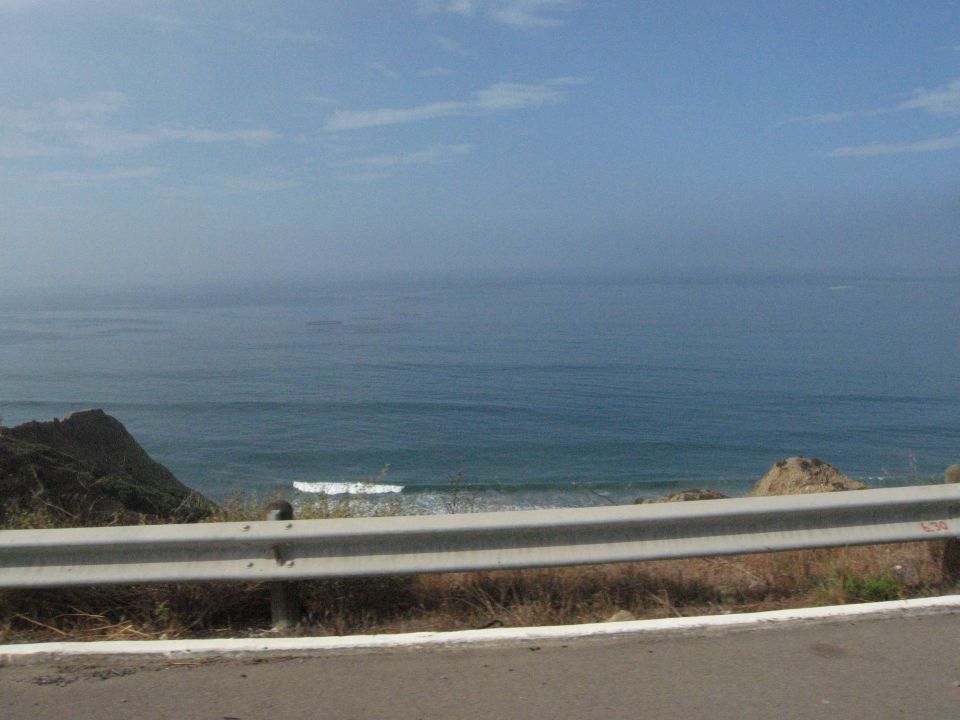 Ocean waves crash near a rocky coastline with a guardrail in the foreground