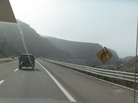 1986 Ford Bronco - A dark SUV travels down a highway alongside a mountainous landscape, with a curve warning sign visible.