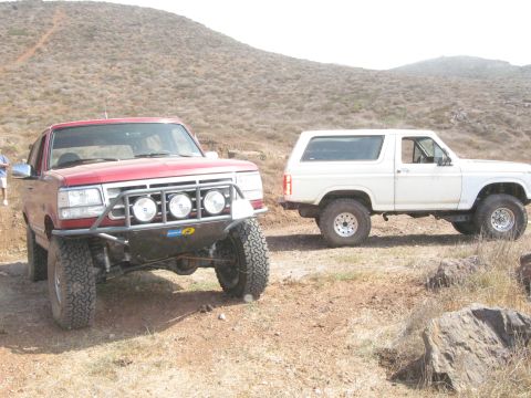1986 Ford Bronco - Two SUVs, a white Ford Bronco and a red custom off-roader, sit parked on a dusty, arid hillside.