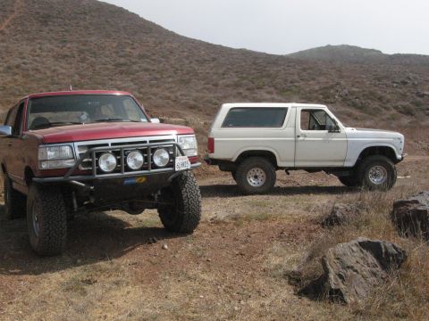 1986 Ford Bronco - Two Ford Broncos parked on a dirt road with hills in the background.