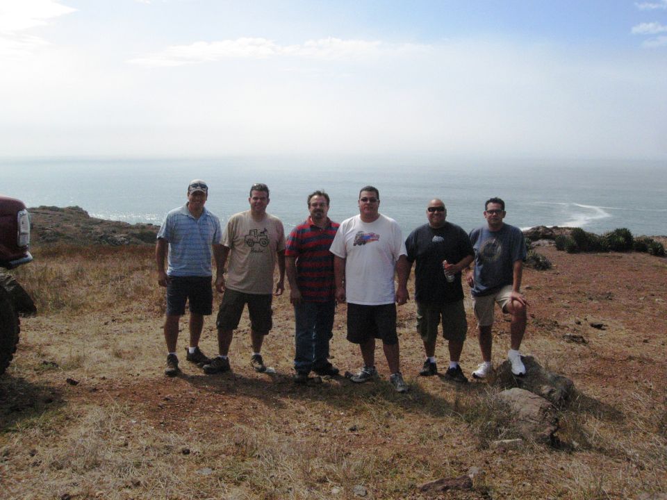Red vehicle fender and tire on the left, six men stand on a dirt hill overlooking a hazy ocean