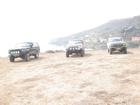 1986 Ford Bronco - Three modified SUVs are parked on a dusty bluff overlooking a coastal landscape with buildings on the hillside.