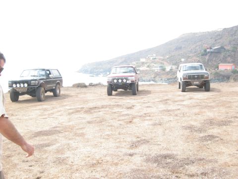 1986 Ford Bronco - Three modified SUVs are parked on a dirt lot overlooking the ocean and a hillside with houses.